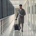 stylish man with suitcase and passport walking along airport corridor